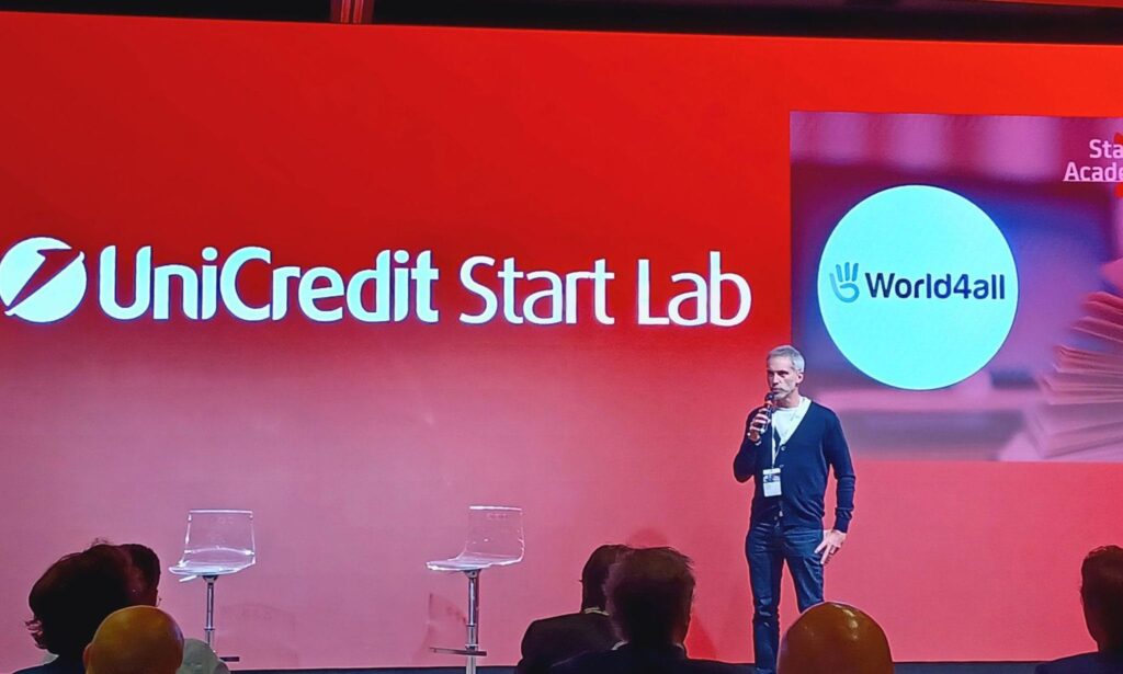 Speaker on a red-lit stage with a large screen showing the UniCredit Start Lab logo and the World4All circle in the background, presenting to an audience.