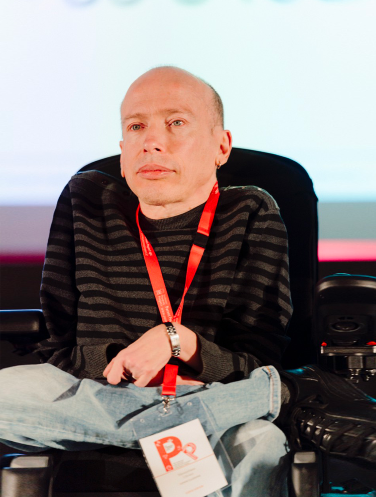 Man with a shaved head wearing a black and gray striped sweater and a red lanyard, seated on a stage in a wheelchair with a badge hanging from the lanyard.
